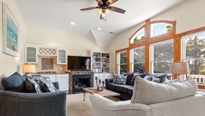 A spacious living room with a fireplace, a television, and a large window offering a view of the outdoors. - Ocean Escape: Lakefront, Pool, Hot Tub, Game Room (Corolla)