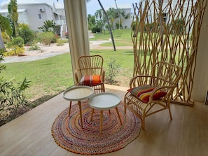 A patio with wicker chairs and a round table, overlooking a well-maintained garden and a white building in the background.