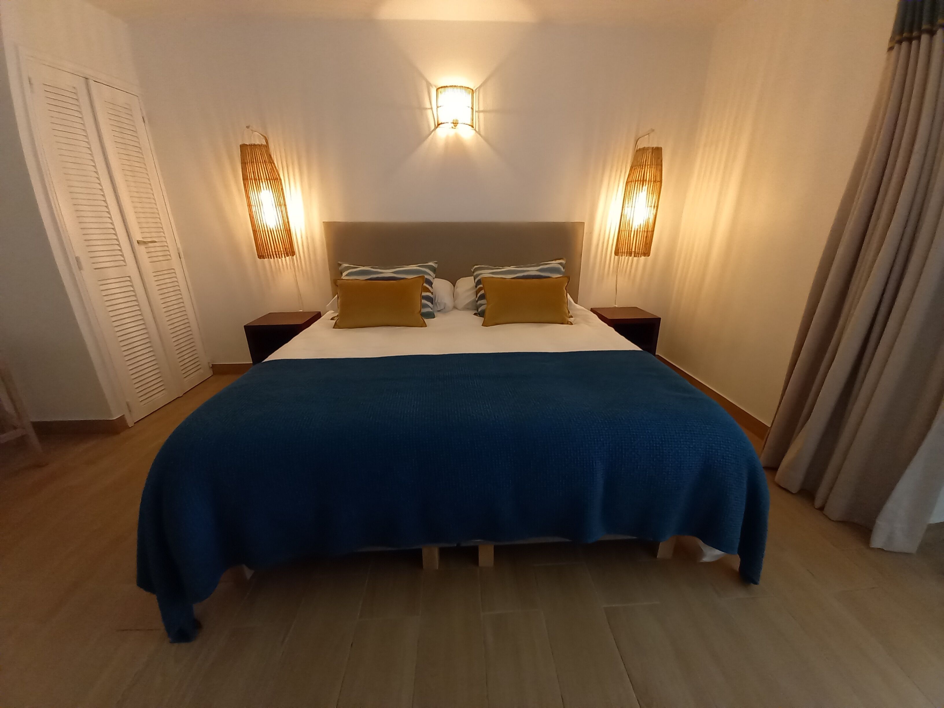 Un dormitorio con una cama grande, dos mesitas de noche, una lámpara fijada a la pared y una puerta con contraventanas.