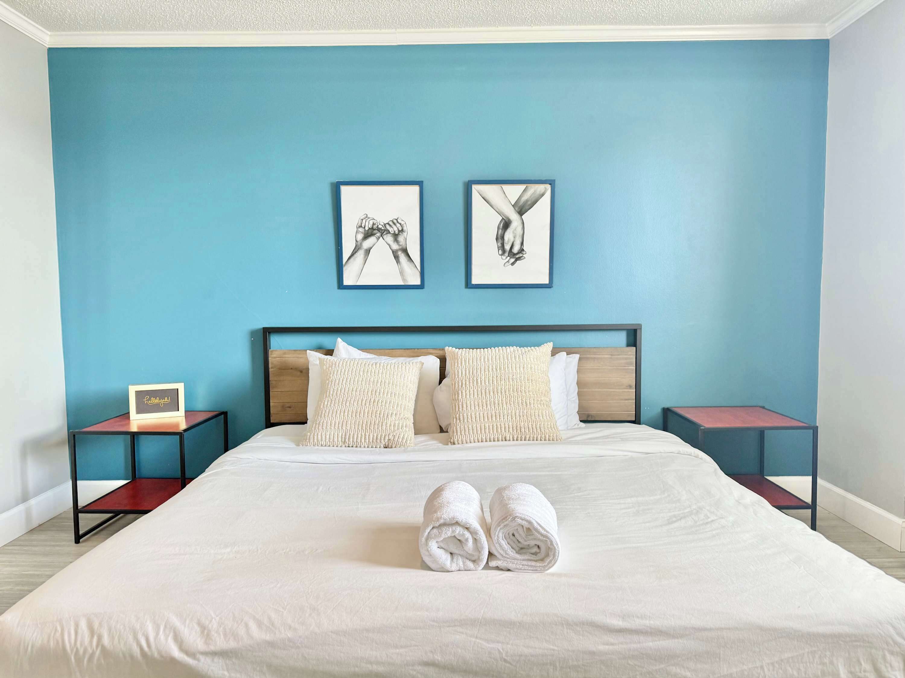 A hotel room with a bed, two pillows, two nightstands, two framed artworks, and two rolled towels on the bed.