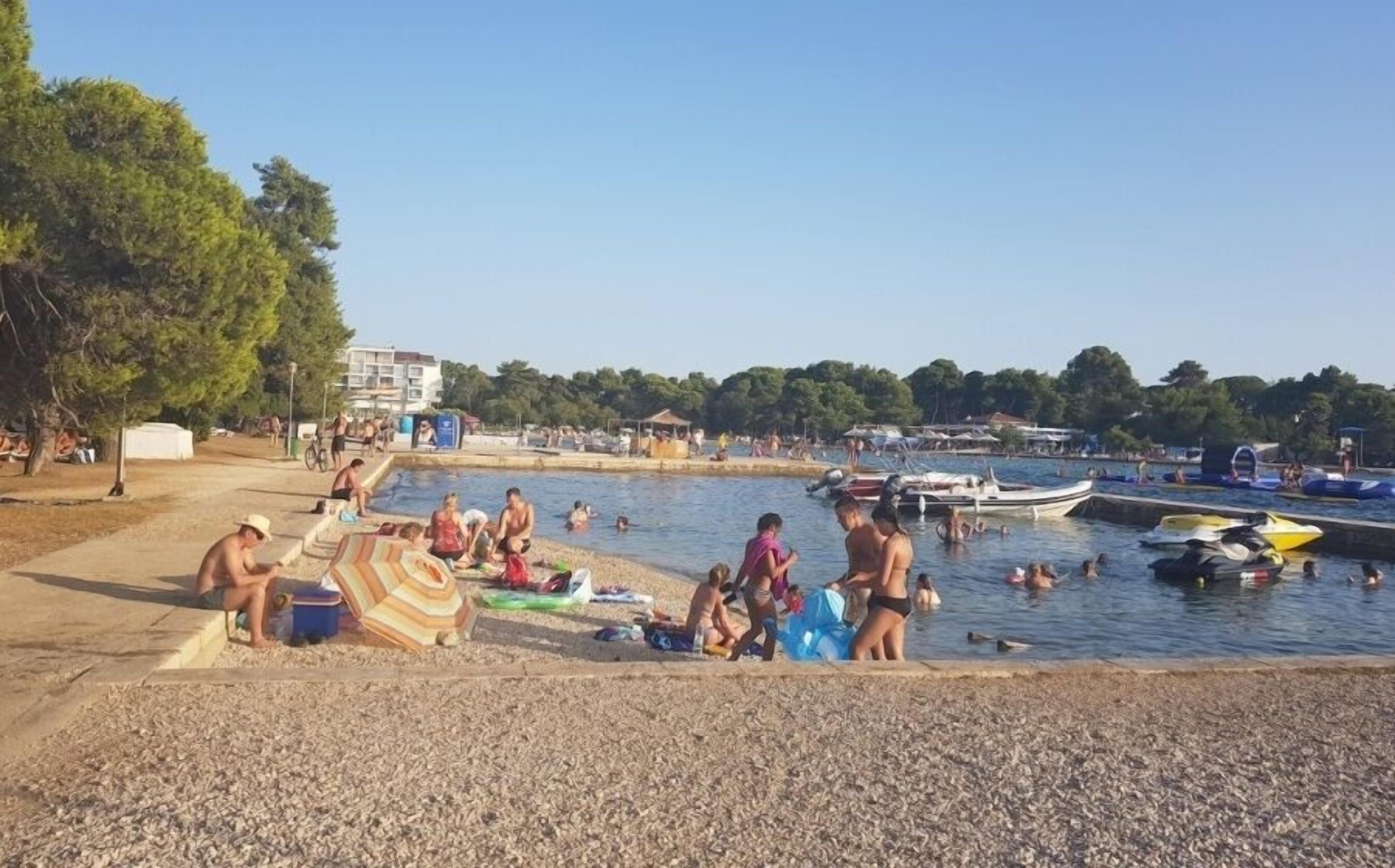 A beach with people swimming and sunbathing, boats in the water, and a building in the background.
