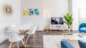 Un salon moderne avec une table à manger blanche, des chaises blanches, un fauteuil bleu, un téléviseur, une plante et des œuvres d’art accrochées au mur.