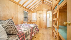 A wooden cabin room with a bed, a window with blinds, a door, and a small bathroom visible through a door. - The Colorado Spruce Cabin #15 (Bayfield)