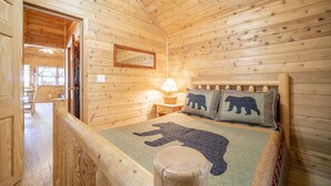 A wooden cabin room with a bed, a nightstand, a lamp, and a window. - The Blue Spruce Cabin #16 (Bayfield)