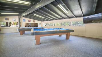 A pool hall with two pool tables, a high ceiling with exposed beams, and windows overlooking a wooded area.
