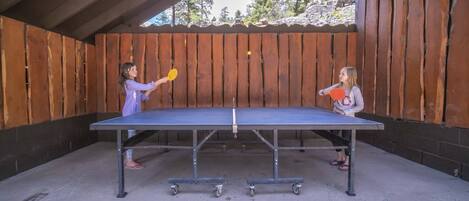An outdoor ping pong table set up in a covered area with wooden walls and a view of trees and rocks in the background.