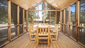 Un espace de restauration extérieur abrité, avec des tables et des chaises en bois, de grandes fenêtres et une vue sur des arbres et un bâtiment en arrière-plan.
