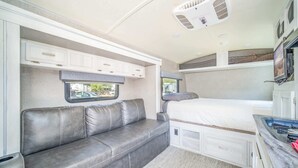 Interior compacto de una caravana con sofá, cama, televisor montado en la pared y pequeña zona de kitchenette.