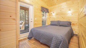 A wooden cabin bedroom with a bed, a door leading outside, and a window with curtains. - The Twilight Cabin #4 (Bayfield)