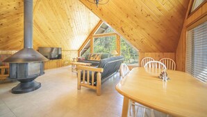 En hyggelig træhytte med synlige hanebånd, pejs, sofa, spisebord og fjernsyn.