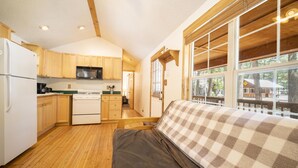 A compact kitchen with wooden cabinets, a white stove, and a microwave. There is a window with a view of trees and a deck outside. - The Iris Cabin #1 Sum (Bayfield)
