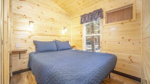 A wooden cabin room with a bed, a window, and a wall-mounted light fixture. - The Fox Den Cabin #14 (Bayfield)