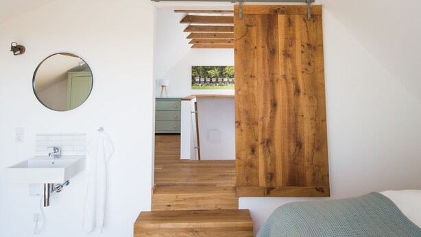 Un dormitorio moderno con escalera de madera, cama, lámpara colgante y baño con espejo redondo y lavamanos.