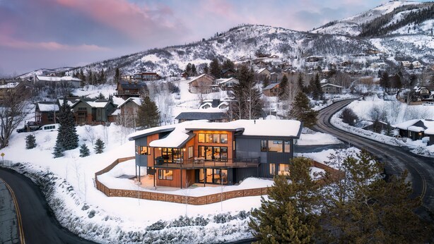A modern two-story house with a circular deck, surrounded by snow-covered trees and a mountainous backdrop.