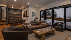 A cozy living room with a leather sofa, a wooden coffee table, and a foosball table. There is a large window with a view of a snowy landscape.