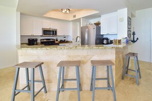 Una cocina con gabinetes blancos, un refrigerador de acero inoxidable, un microondas y una cafetera. Hay tres taburetes frente a la isla de la cocina.