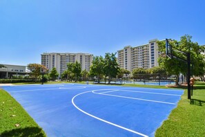 Una cancha de baloncesto al aire libre con piso azul, rodeada de césped y árboles, con edificios altos al fondo.
