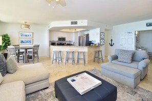 A modern living room with a grey sofa, a black ottoman, and a dining area with a table and chairs. - 5th Floor 3 Bedroom Condo with Semi-Private Elevator (Destin)