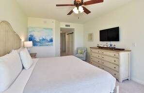 Un dormitorio con cama, cómoda, ventilador de techo, un cuadro, televisor y una puerta que da a otra habitación.