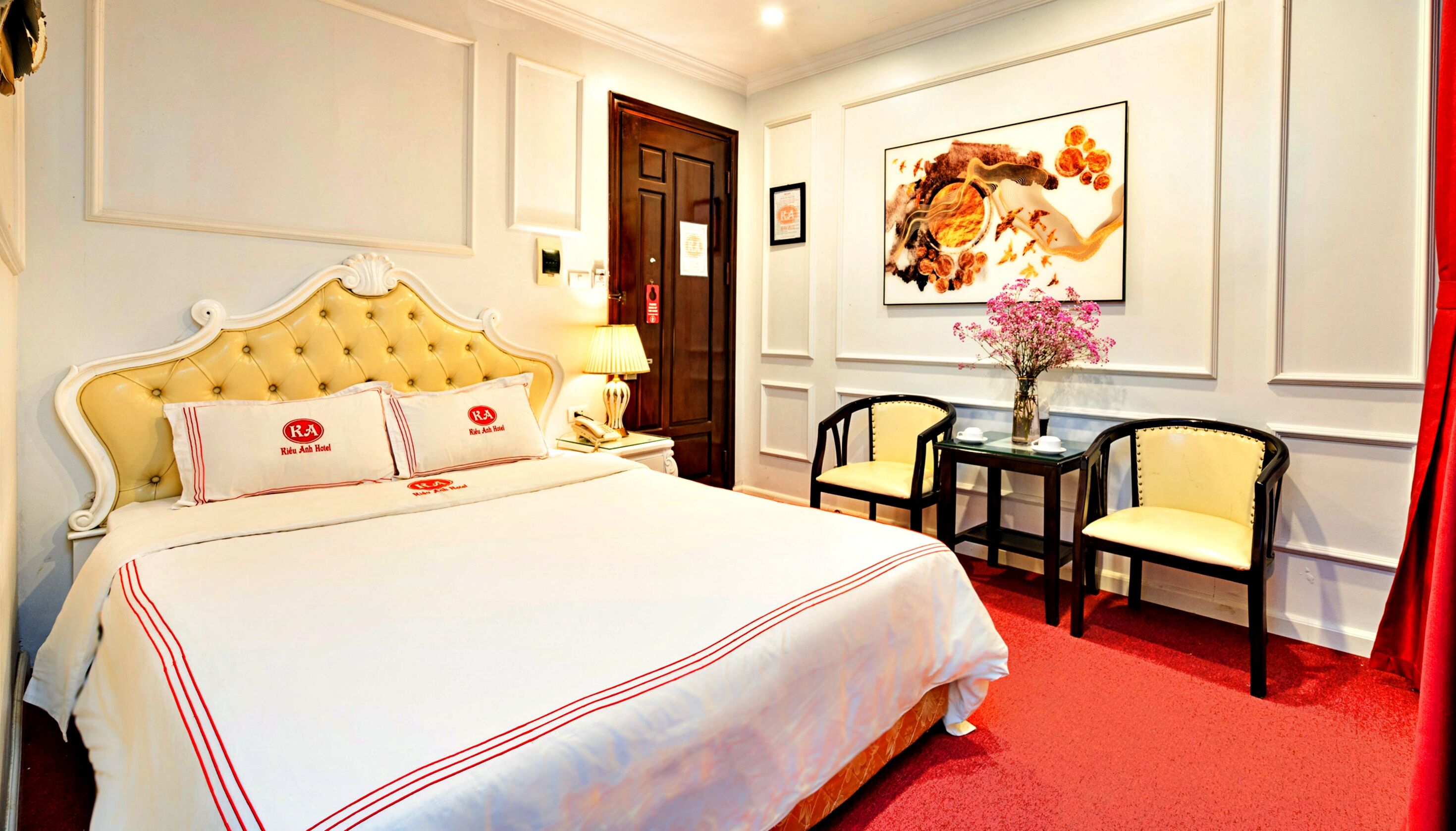 A hotel room with a large bed, a nightstand, a lamp, two chairs, and a painting on the wall.