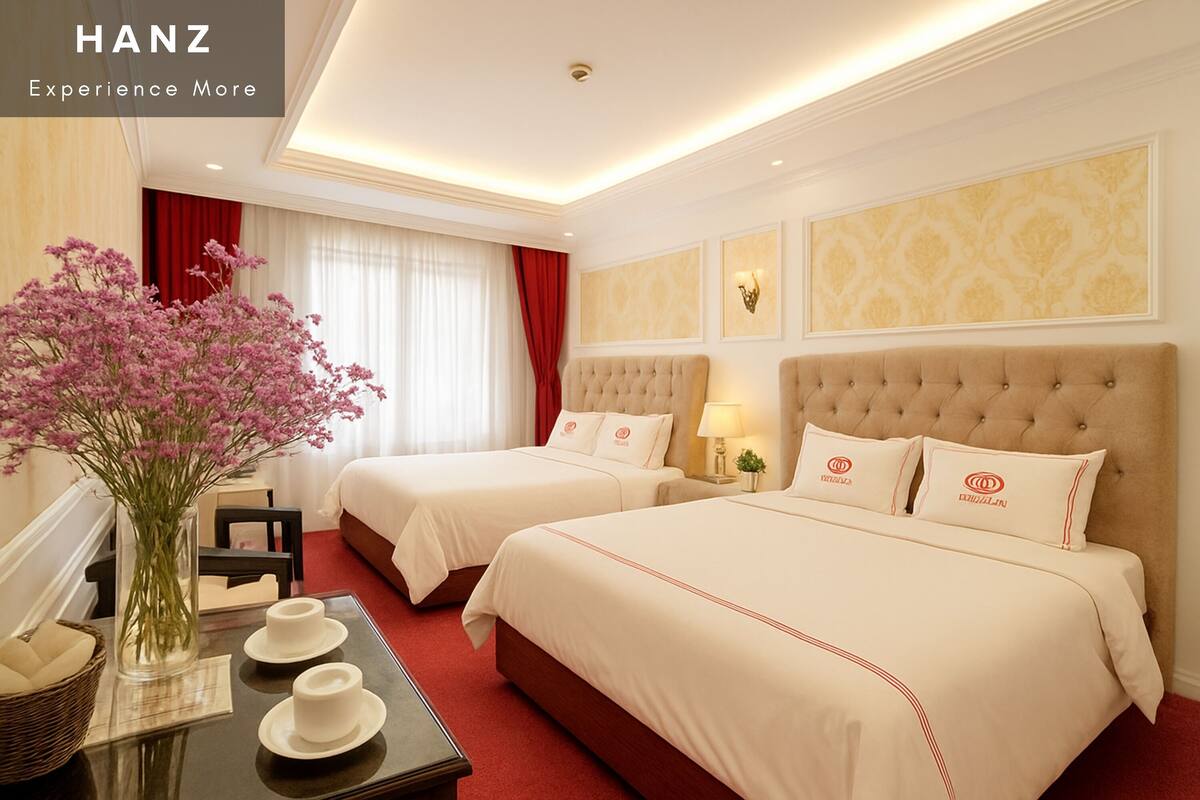 A hotel room with two beds, a vase of flowers, a table with cups, and a red curtain.