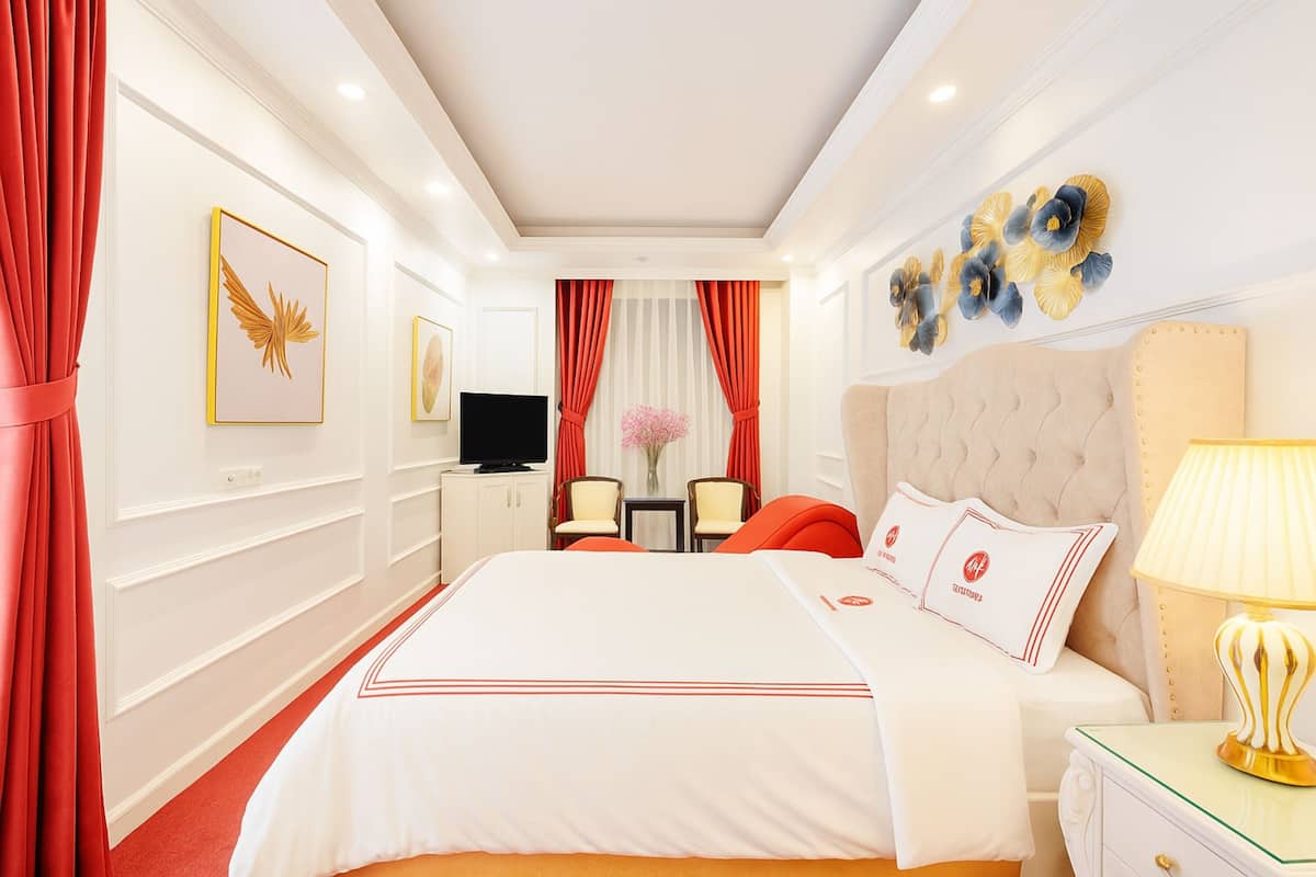 A hotel room with a large bed, red curtains, a flat-screen TV, a small table with chairs, and decorative wall art.