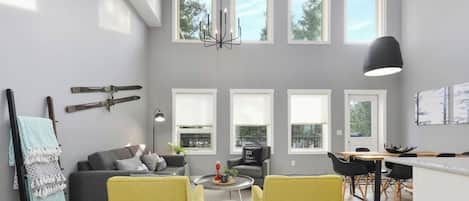 Una sala de estar moderna con un sofá gris, sillas amarillas, un comedor con mesa y sillas, y ventanas amplias con persianas blancas.
