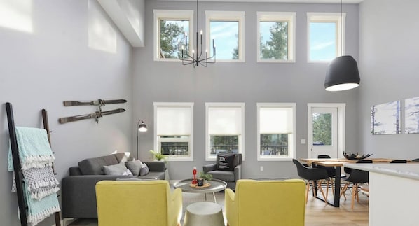 Uma sala de estar moderna com sofá cinza, cadeiras amarelas, área de jantar com mesa e cadeiras, e janelas amplas com persianas brancas.