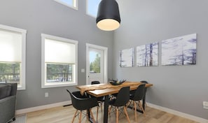 Uma área de refeições com uma mesa de madeira, cadeiras pretas e uma luminária pendente preta. Há três janelas com persianas brancas e uma parede cinza com três quadros emoldurados.