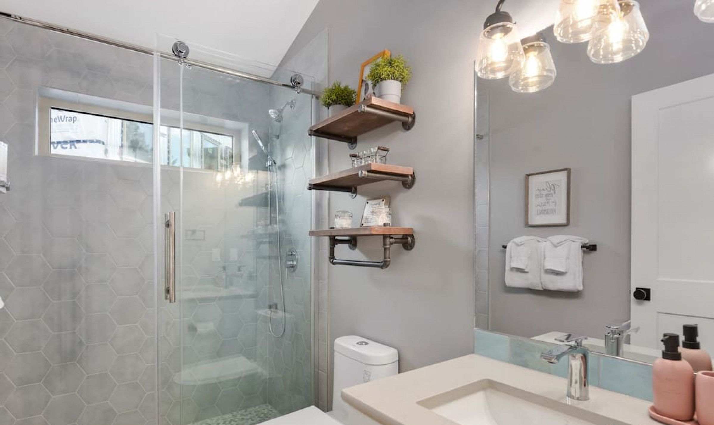 Une salle de bain moderne avec une douche à parois de verre, des toilettes blanches, un lavabo beige et des étagères en bois ornées d’objets décoratifs.