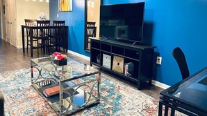 A living room with a blue wall, a glass coffee table, a TV on a black stand, and a dining area with chairs and a table. - Cozy Beautiful 2BD Apt in Center City (Philadelphia)