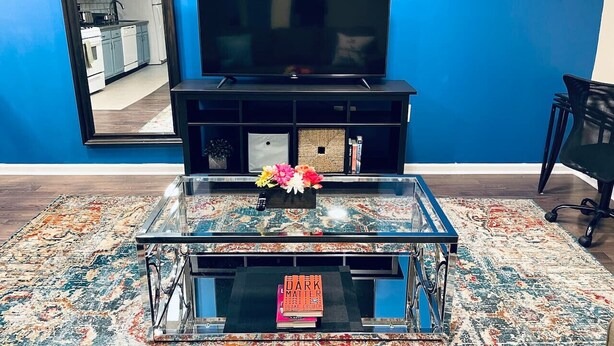 A living room with a blue wall, a flat-screen TV, a glass coffee table with a book, and a patterned rug. - Cozy Beautiful 2BD Apt in Center City (Philadelphia)