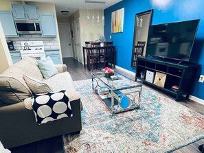 Ein Wohnzimmer mit einer Couch, einem Couchtisch und einem Fernseher. Im Hintergrund ist eine Küche zu sehen.