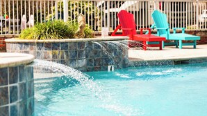 A swimming pool with a water feature, surrounded by a fence and red and blue lounge chairs. - Sugar Sands RV Resort (Gulf Shores)