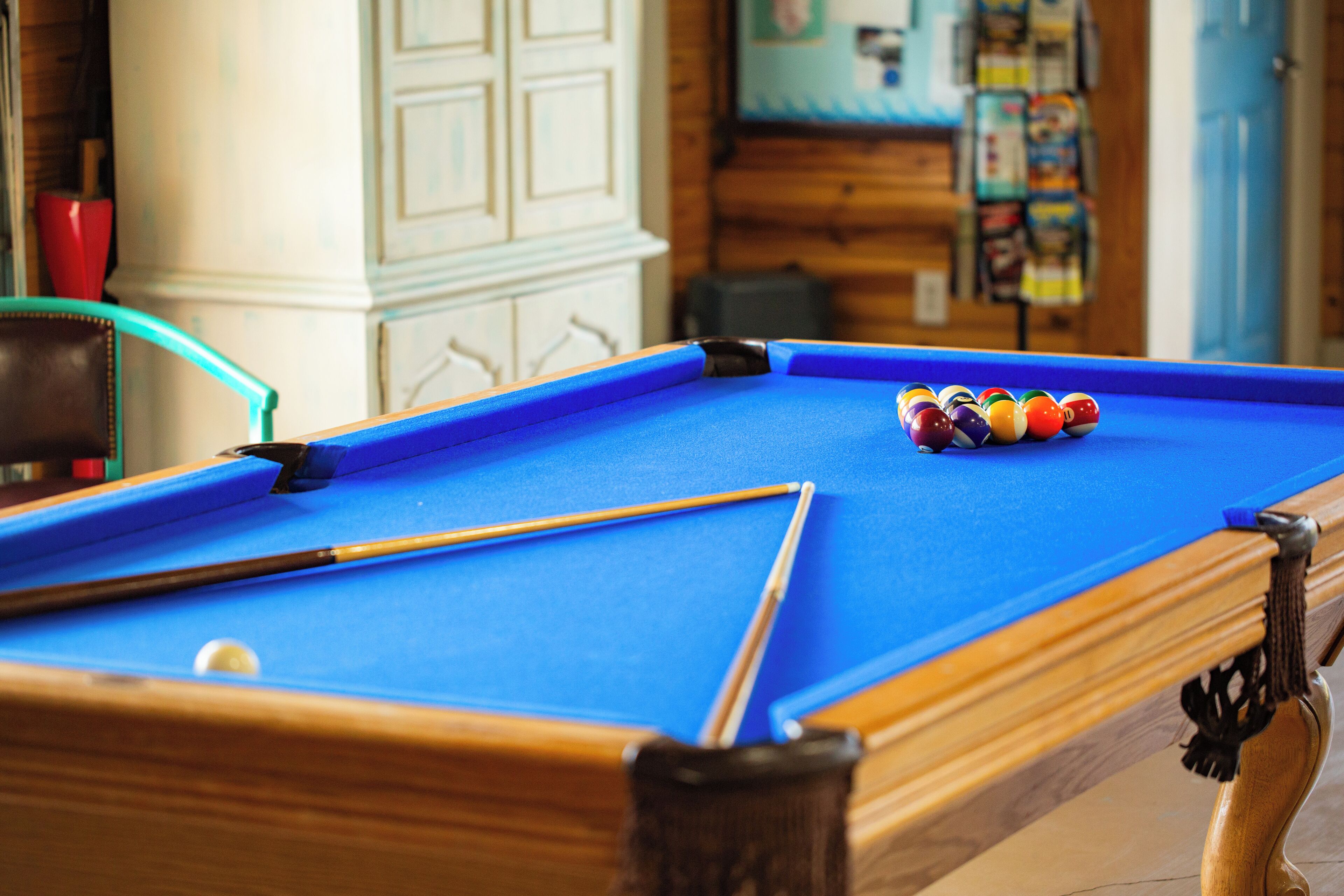 A pool table with balls and cues, a wooden chair, and a cabinet in the background.