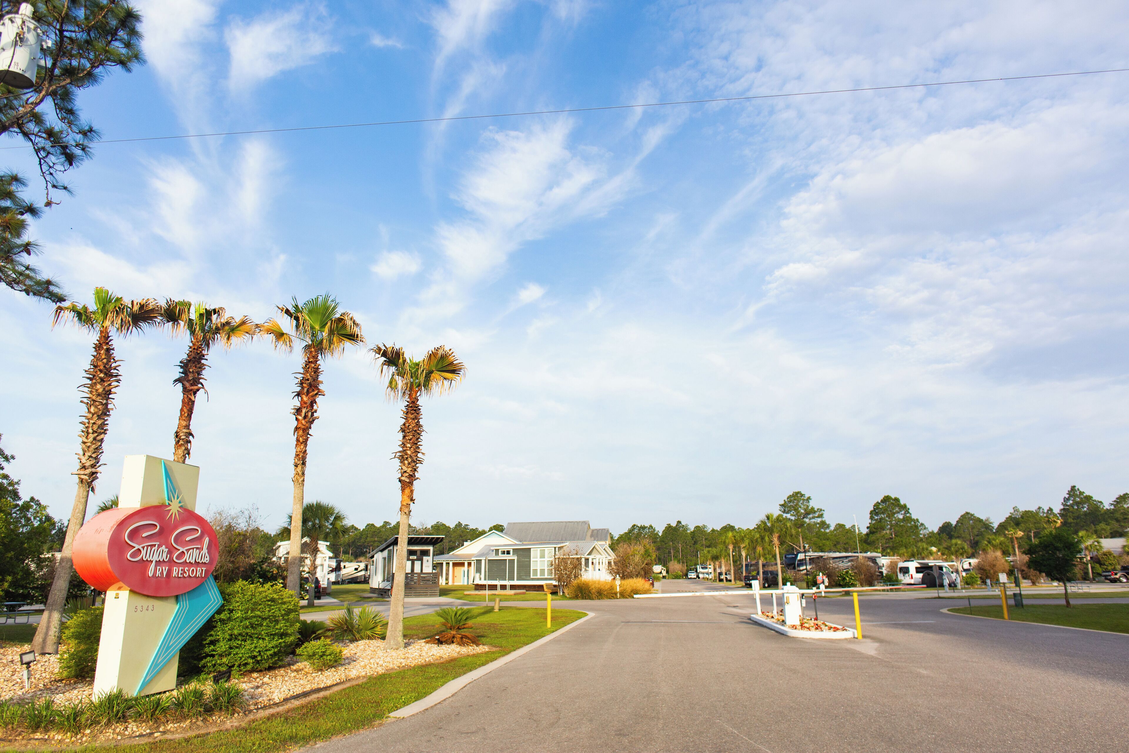 A road leading to a resort with a sign for Sugar Sands URY Resort, surrounded by palm trees and a clear sky.