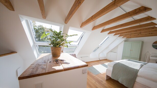 Une chambre avec un plafond en bois, des fenêtres de toit, un lit, une table de chevet et une plante verte.