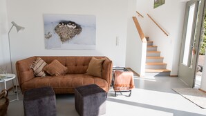 Un salon moderne avec un canapé marron, deux poufs, un tableau représentant des hiboux, un escalier et une grande fenêtre.