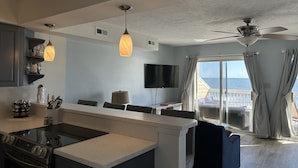 A modern kitchen with a central island, pendant lights, a dining area with a TV, and a balcony with a view of the sea. - Beach Front Property with Stunning  Ocean Front View in the Ocean Dunes Resort (Kure Beach)