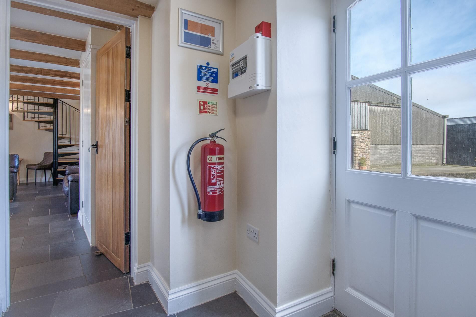 A hallway with a fire extinguisher mounted on the wall, a fire action sign, and a window with a view of a building outside.