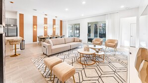 Una sala de estar moderna con un sofá, sillones y una mesa de centro. Cuenta con un comedor con mesa y sillas. La habitación tiene ventanas grandes y una zona de cocina con refrigerador.