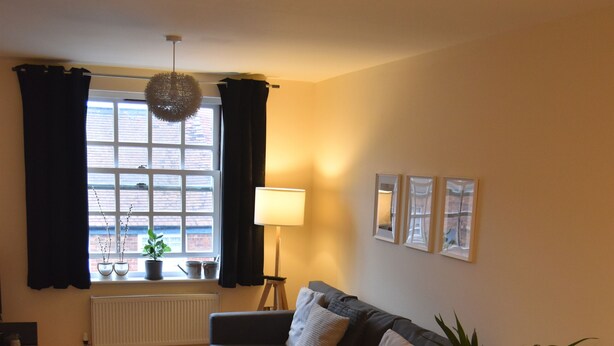 A modern living room with a sofa, a small round table with a plant, a lamp, and a view of the outside through a window with curtains. - Scandinavian City Centre Apartment (Worcester)