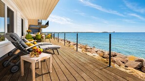 Una terraza de madera con mobiliario exterior, vista al mar y un edificio con ventanas amplias.