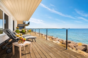 A wooden deck with outdoor seating, a view of the sea, and a building with large windows.