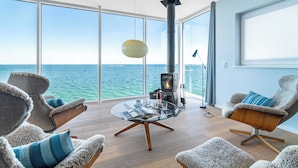 Una sala de estar moderna con una mesa de vidrio, sillas cómodas, una chimenea y un ventanal grande con vista al mar.