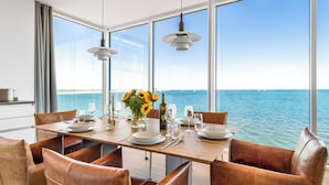 Un comedor con vista al mar, una mesa de madera dispuesta con platos, vasos y una botella, y sillas de cuero.