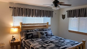Une chambre avec un lit en bois, un ventilateur de plafond, une lampe et une fenêtre avec des stores.