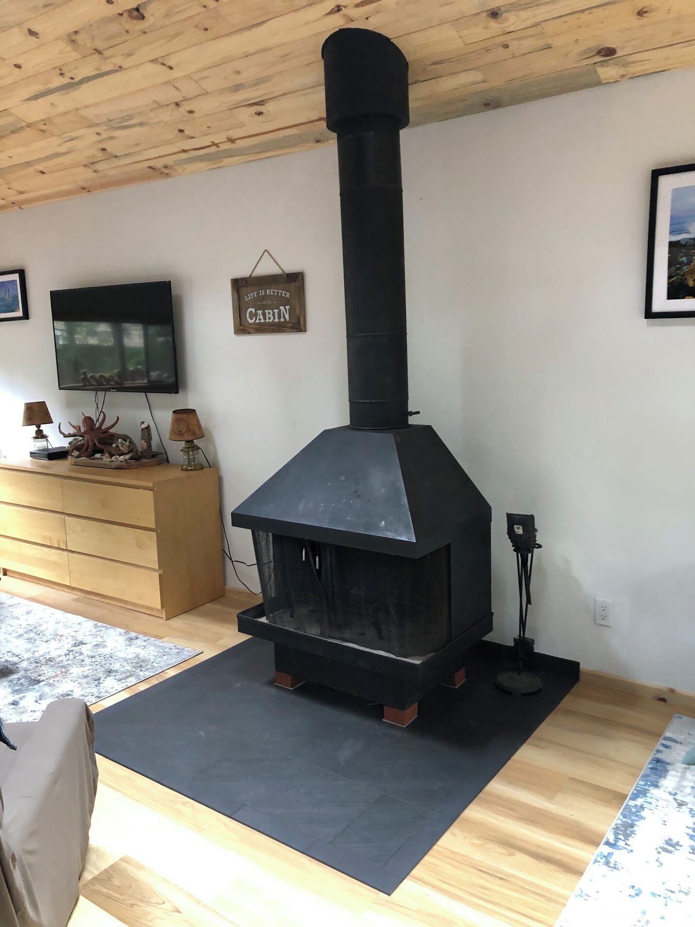 A living room with a black wood-burning stove, wooden flooring, a television, and a sign that reads 'Life is better cabin.'
