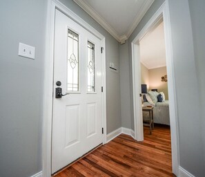 Enter a hallway with a door featuring a geometric glass panel, leading to a cozy room with a bed, table, and lamp.