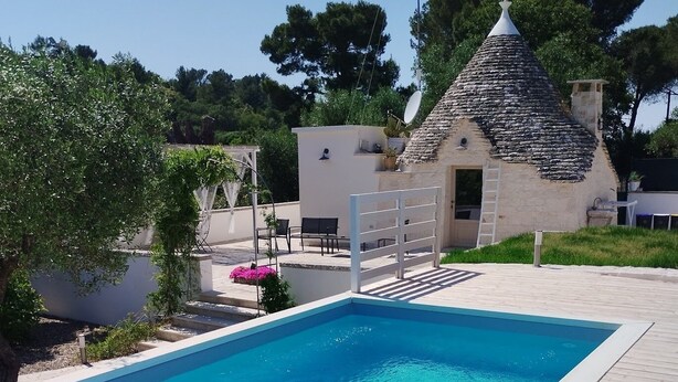 A swimming pool with a wooden deck, a stone-roofed building, and a white wall with a door. - Romantic Trullo with Pool: Entire Private Property❣️ (Alberobello)